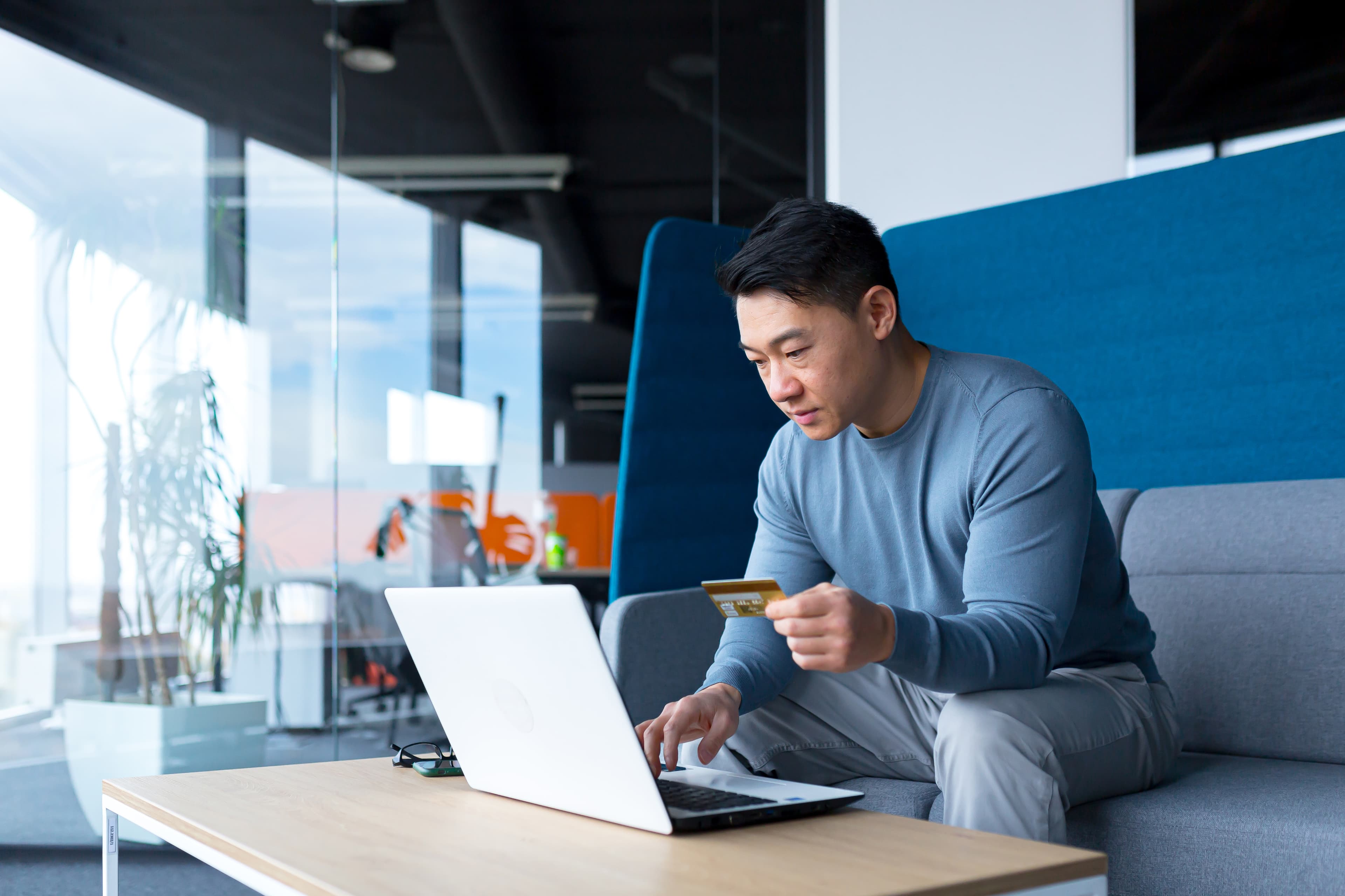 Man sitting on grey sofa looking at laptop while holding credit card